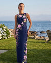 Woman in a floral dress standing by the ocean with flowers and a chair in the background.